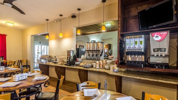 A café with multiple seating areas, a coffee bar with various drink dispensers, and a television mounted on the wall. - Clarion Inn & Suites (Mission)