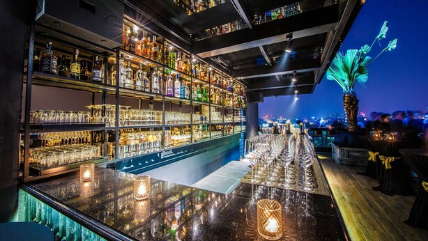 Un espace bar avec des étagères remplies de bouteilles d’alcool et de verres, un comptoir de bar élégant et une vue sur la ville au crépuscule.