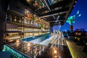 Zona de bar con estantes de botellas de licor y vasos, una barra pulida y vistas a la ciudad al atardecer.