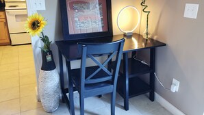 Un pequeño comedor con una mesa, dos sillas, una obra de arte enmarcada, un jarrón con un girasol y una lámpara fijada a la pared.