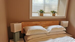 Un dormitorio con cabecera de madera, ropa de cama blanca, una mesita de noche con lámpara y una ventana con persianas y dos plantas en macetas.