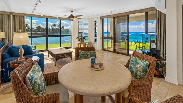 A spacious living area with a large window overlooking the ocean, furnished with wicker chairs, a round table, and a blue sofa. - Kuhio Shores Oceanfront Corner Unit (Koloa)