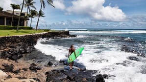 A person with a surfboard on a rocky beach with palm trees and a building in the background. - Kuhio Shores Oceanfront Corner Unit (Koloa)