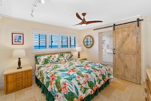 A bedroom with a bed, two bedside tables, a ceiling fan, a window with shutters, and a wooden door.