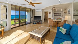 A spacious living area with a large TV, a blue sofa, and a balcony with a view of the ocean. - Kuhio Shores Oceanfront Corner Unit (Koloa)