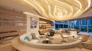 A modern lounge area with a curved sofa, armchairs, and a central coffee table. There are bookshelves with framed pictures and a unique ceiling light fixture.