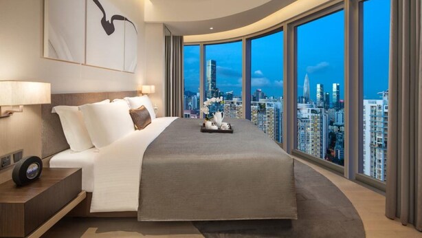 A modern hotel room with a curved bed, a digital clock, and a city view through large windows.