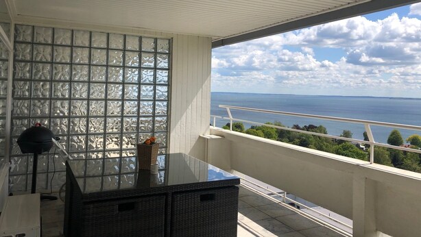 A balcony with a view of the sea, a glass table, wicker furniture, and a decorative vase.