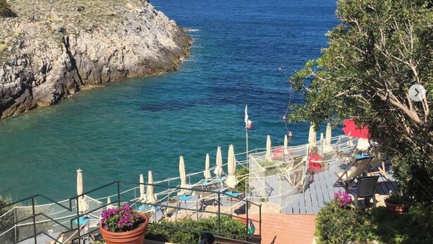 A coastal view with a rocky cliff, a clear blue sea, and a terrace with outdoor seating and umbrellas.