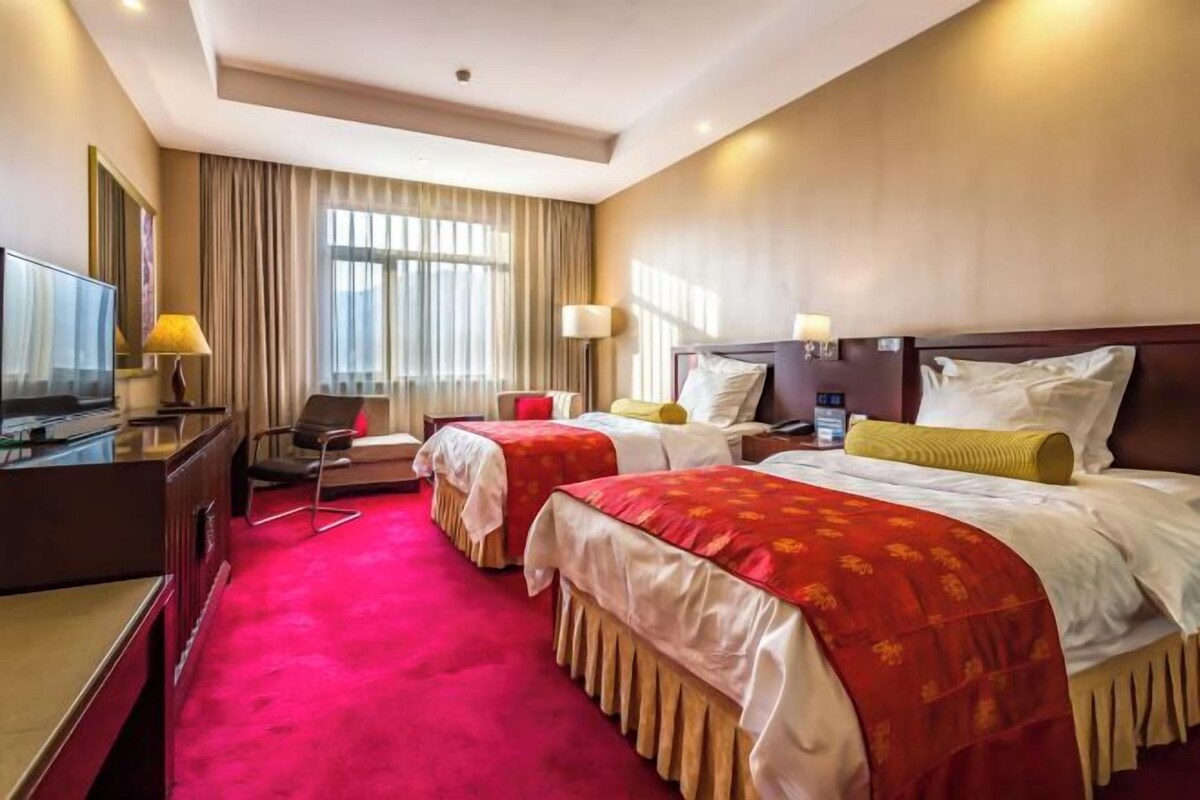 A hotel room with two beds, a desk with a TV, and a large window with curtains.