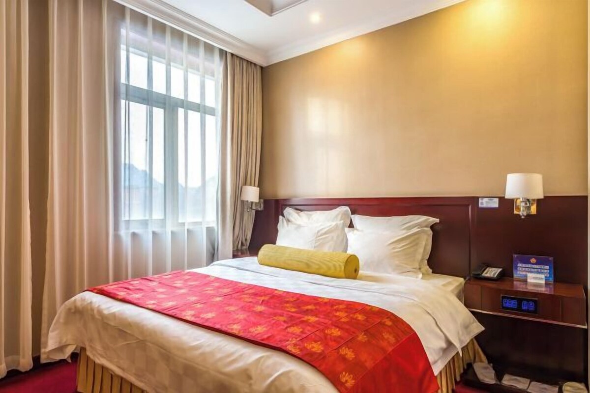 A hotel room with a large bed, a wooden headboard, a bedside table with a lamp, and a view of the outdoors through the window.