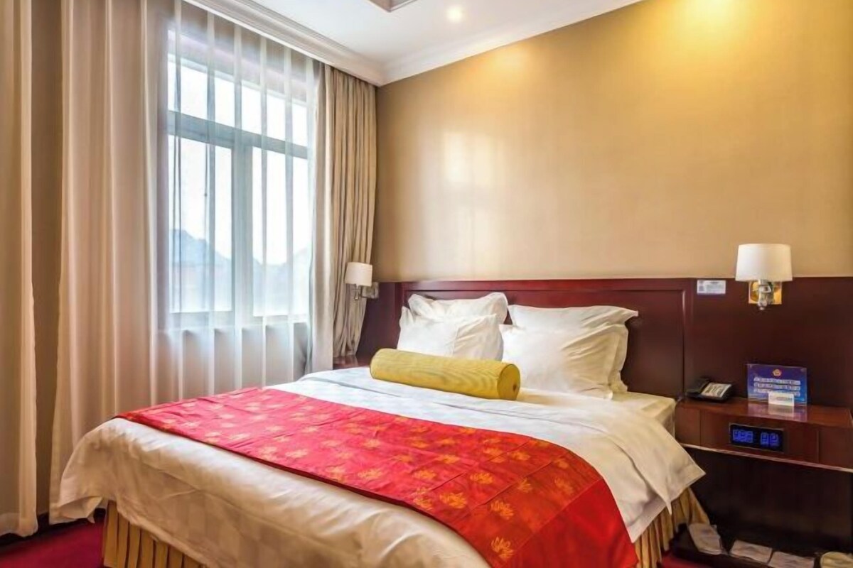 Una camera d'albergo con un letto grande, una testiera in legno, un comodino con una lampada e vista sull'esterno dalla finestra.
