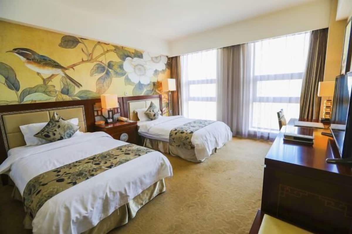 A hotel room with two beds, a desk, a television, and a large window with curtains.