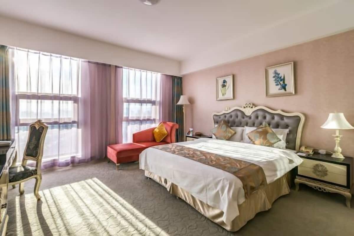 A hotel room with a large bed, a red armchair, a desk with a chair, and a window with curtains.
