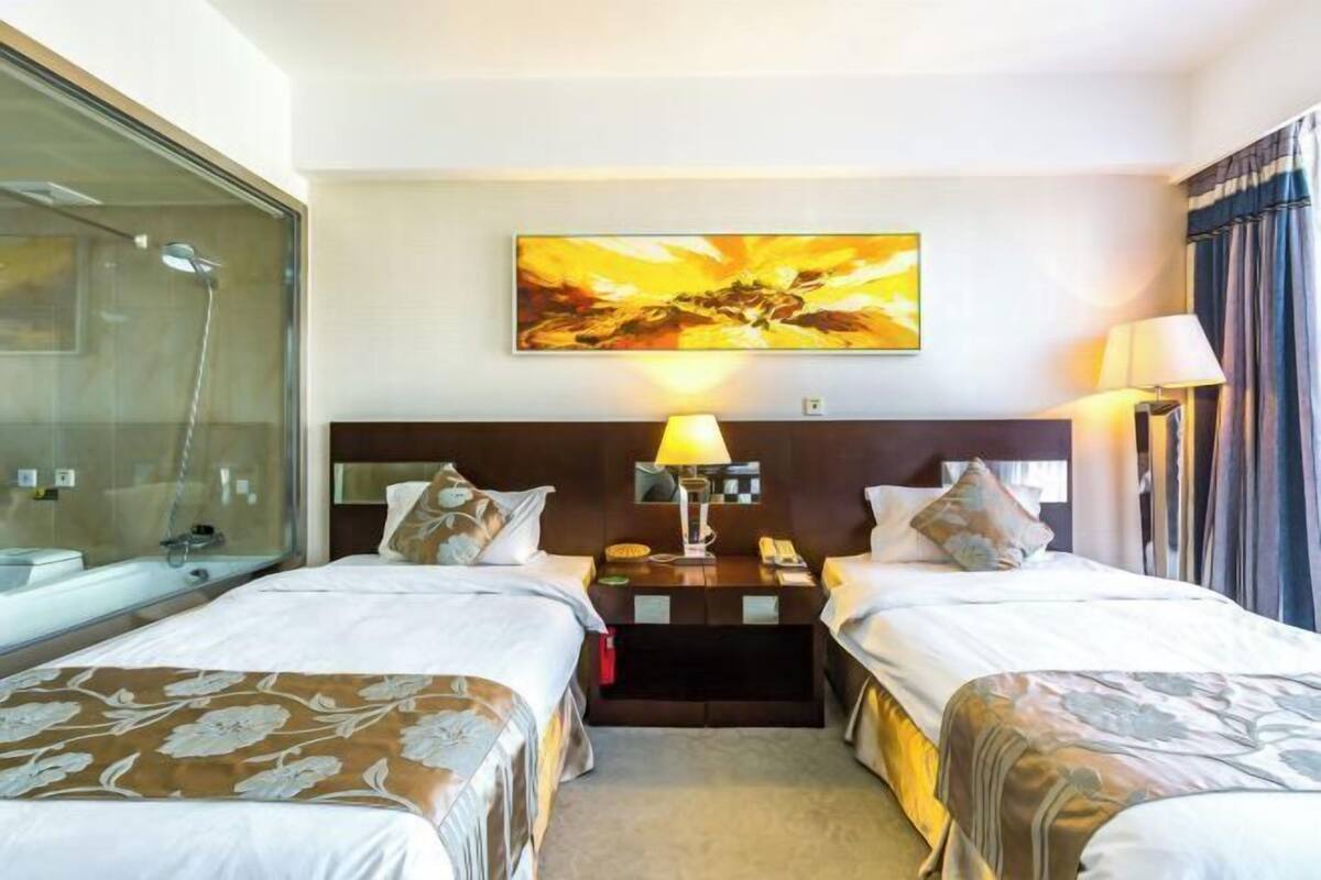 A hotel room with two beds, a large window, a shower, and a painting on the wall.