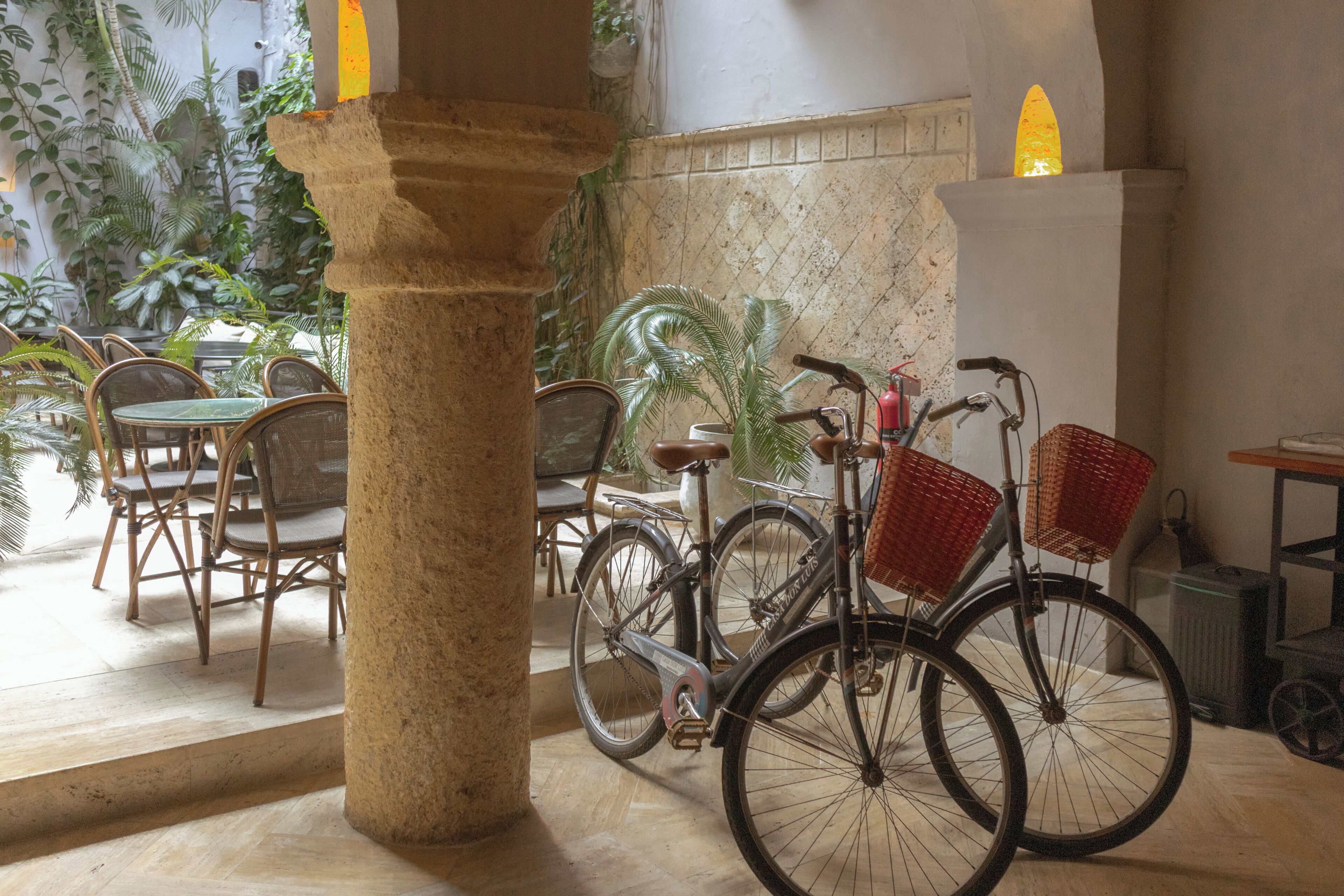 Une pièce avec une colonne en pierre, des chaises en osier, un vélo avec des paniers et un mur carrelé avec une fenêtre.