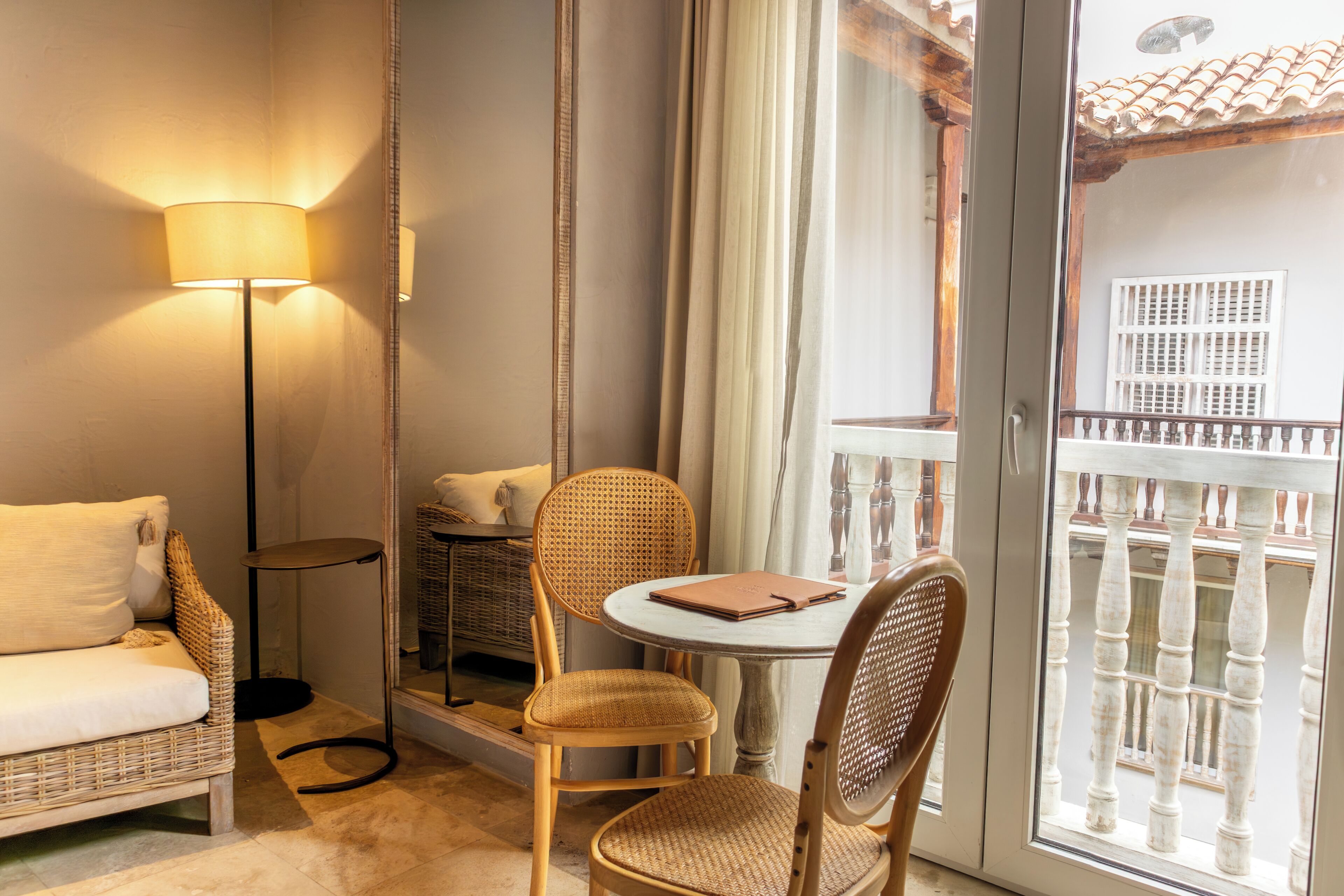 A room with a wicker sofa, a round table with a notebook, a floor lamp, and a balcony with a railing.