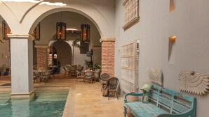 Un espace piscine agrémenté d’un banc, de chaises et de tables, entouré d’arcades et d’un mur en briques.