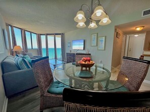 Ein Wohnzimmer mit einem Glastisch, Korbstühlen, einer Couch, einem Fernseher und Blick aufs Meer.