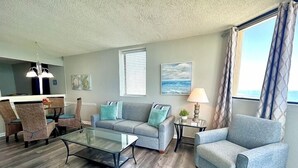 A living room with a glass coffee table, a gray sofa, a blue armchair, and a dining area with chairs and a chandelier.