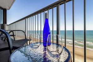 Ein Glastisch mit zwei leeren Weingläsern und einer blauen Flasche auf einem Balkon mit Blick auf den Strand.
