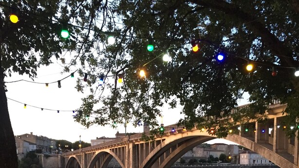 Zona de comedor al aire libre con mesas y sillas bajo un puente, con vistas a un río.