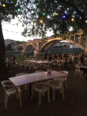 An outdoor dining area with tables and chairs under a bridge, overlooking a river.
