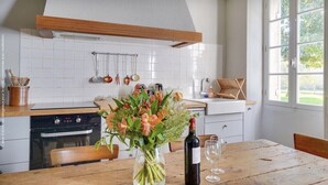 Una cocina con baldosas blancas, vigas de madera, una mesa de comedor con sillas, un jarrón de flores, una botella de vino y una ventana con vistas al verdor.