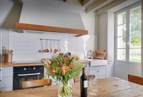 A kitchen with white tiles, wooden beams, a dining table with chairs, a vase of flowers, a wine bottle, and a window with a view of greenery.