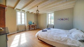 Un dormitorio con cama, piso de madera, dos ventanas, un escritorio y una lámpara de araña.