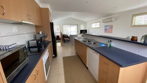 A modern kitchen with a toaster, coffee maker, and dishwasher. There is a dining area with a table and chairs. - Perfect Location - Inverloch (INVERLOCH)