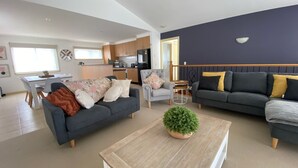 A modern living room with a grey sofa, a smaller armchair, and a coffee table with a green plant. A kitchen is visible in the background with wooden cabinets and a dining area. - Perfect Location - Inverloch (INVERLOCH)