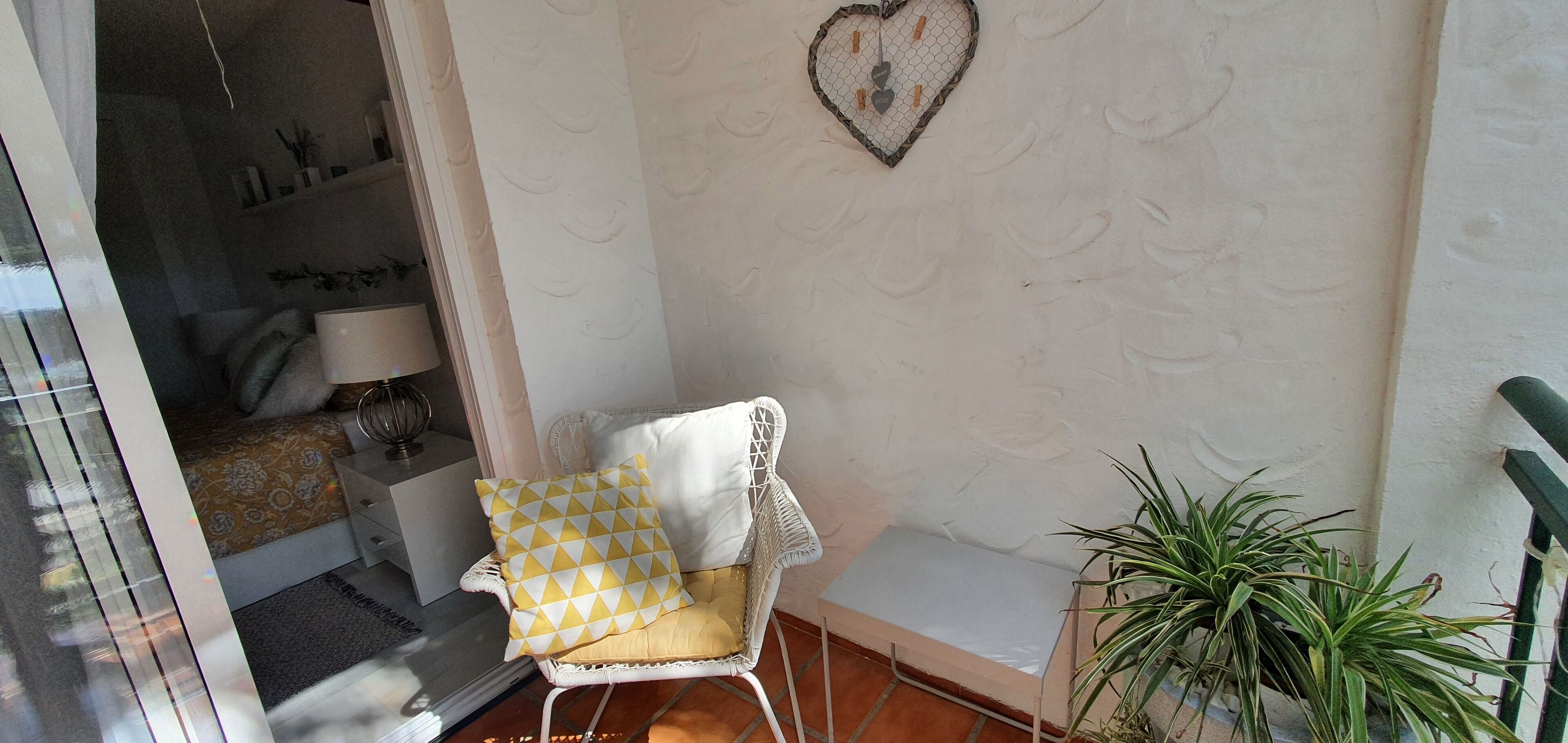A corner of a room with a white wicker chair with a yellow and white cushion, a heart-shaped wall decoration, and a potted plant.