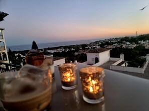 Una mesa con tres velas encendidas y un cuenco, con vistas a un pueblo costero al atardecer.