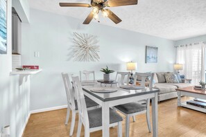A dining area with a white table and chairs, a ceiling fan, and a decorative wall piece. - Sandy Shore: Oceanfront Condo Walking Distance to Golf Course (Caswell Beach)