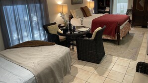 A hotel room with two beds, a dining table, wicker chairs, a bookshelf, and a window with curtains. - Quiet Studio, minutes from Beaches, Shopping, Dining, 5th Ave and 3rd Street (Naples)