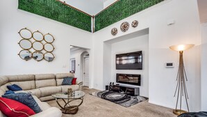 A modern living room with a beige sofa, a glass coffee table, and a red accent chair. A flat-screen TV is mounted on the wall, and a floor lamp is visible. - Beautiful 2 story luxury home in an upscale area! (Houston)