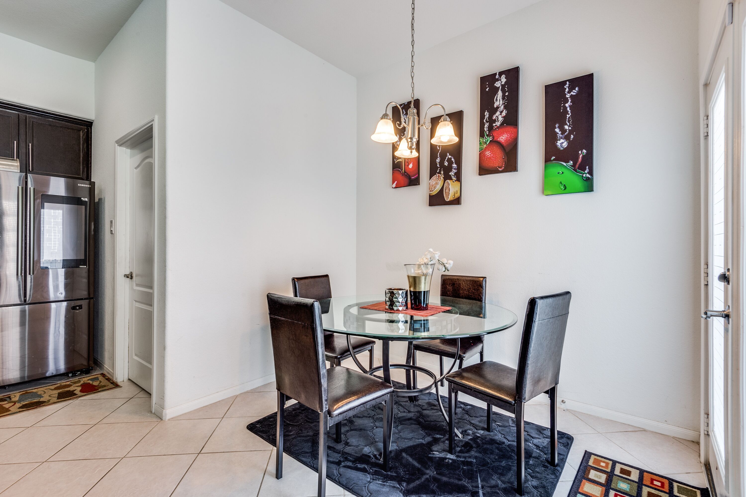 Une cuisine moderne avec un réfrigérateur en acier inoxydable, une table à manger en verre et quatre chaises, ainsi qu’un tapis noir. On y trouve trois luminaires fixés au mur et deux tableaux sur le thème des fruits.