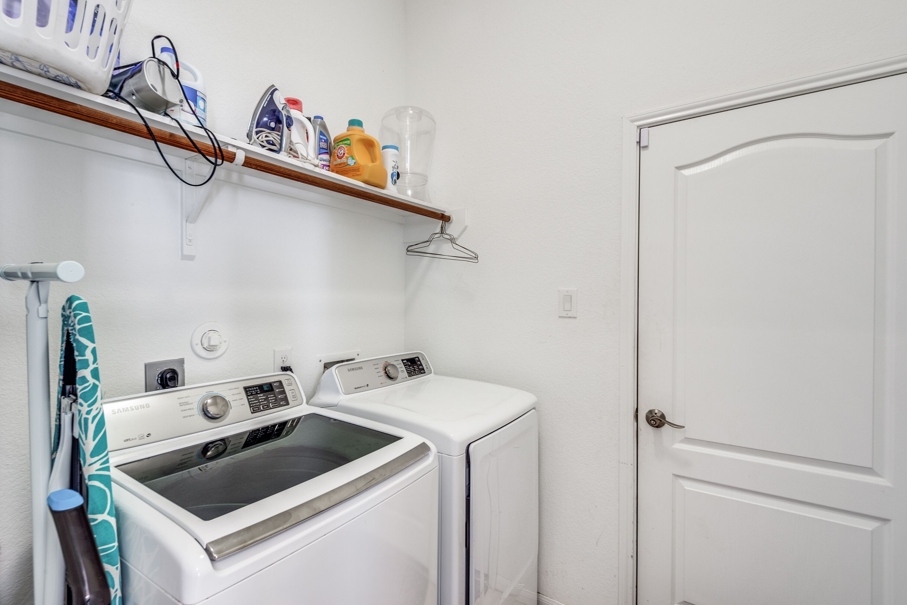 Una sala de bugaderia amb dues rentadores i assecadores blanques, una prestatgeria amb productes de neteja i una porta a la dreta.