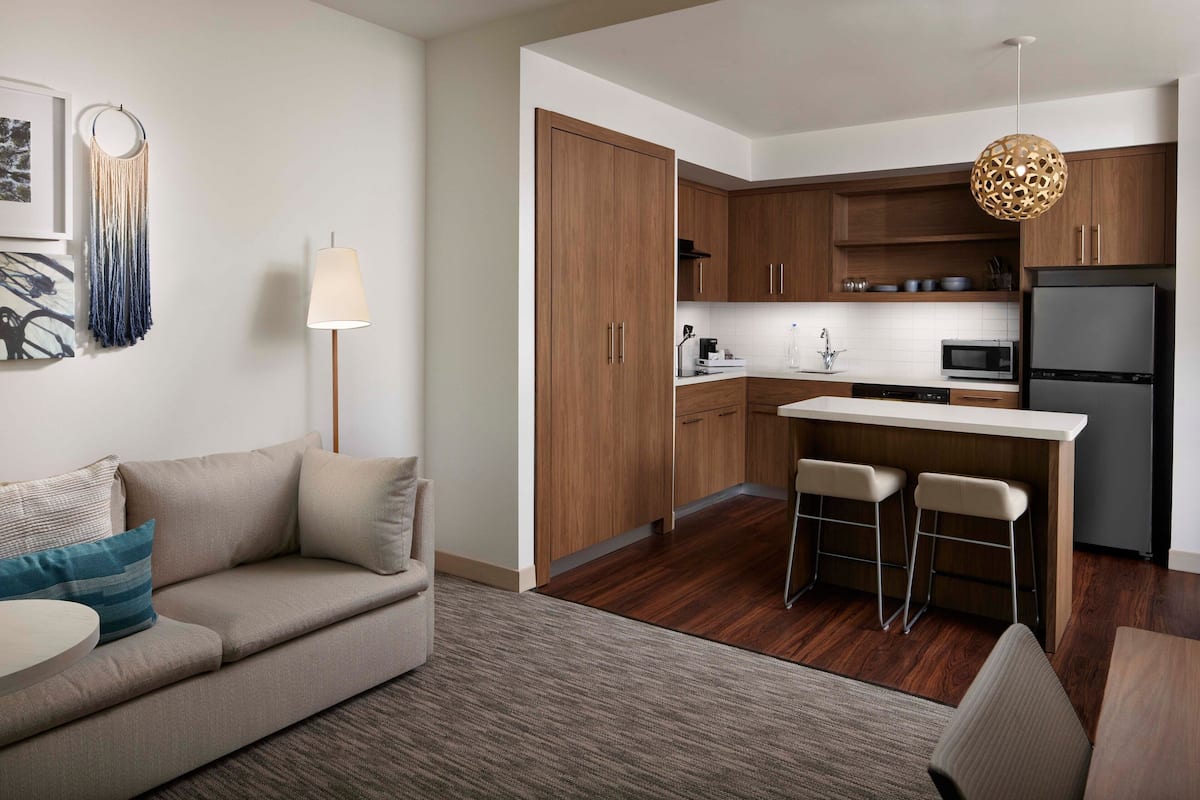 A modern kitchen with wooden cabinets, a central island with stools, a refrigerator, and a pendant light.