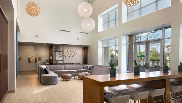 A spacious living area with a large sofa, a wooden coffee table, and a bar with stools. There are large windows providing natural light and a view of the outdoors.