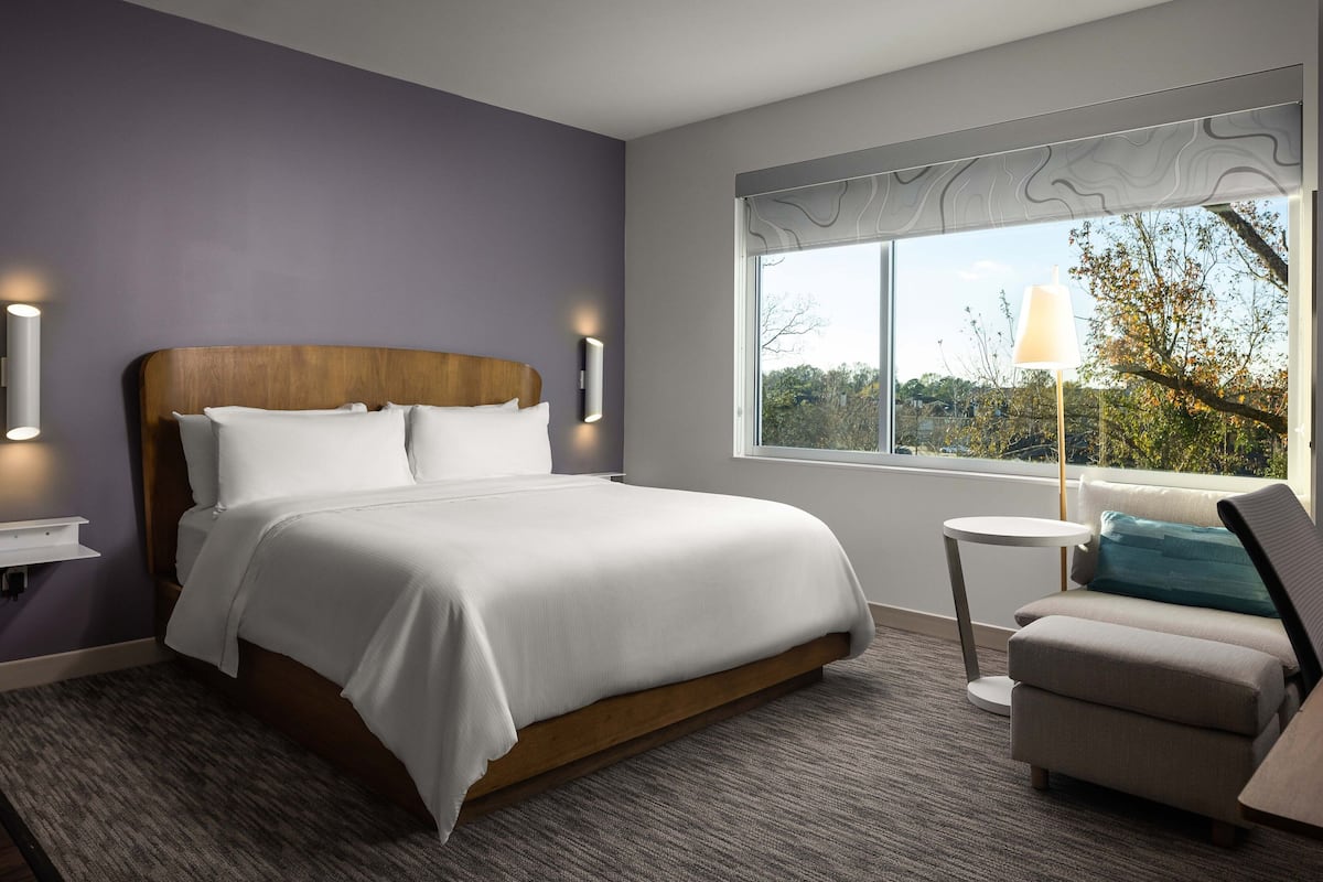 A modern hotel room with a large bed, a small round table, a chair, and a window with a view of trees.