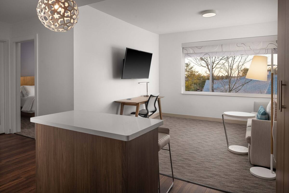 A modern hotel room with a wooden accent wall, a flat-screen TV mounted on the wall, a small round table with a chair, a sofa, and a window with a view of trees.