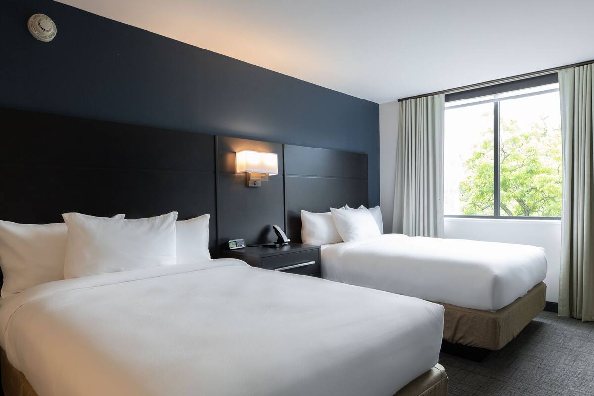 A hotel room with two beds, a nightstand, a lamp, and a window with curtains.