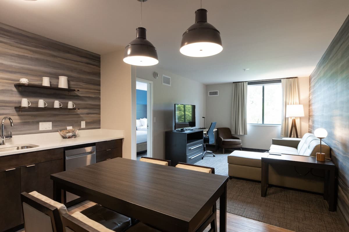 A modern hotel room with a kitchen area, a dining table, a sofa, and a TV.