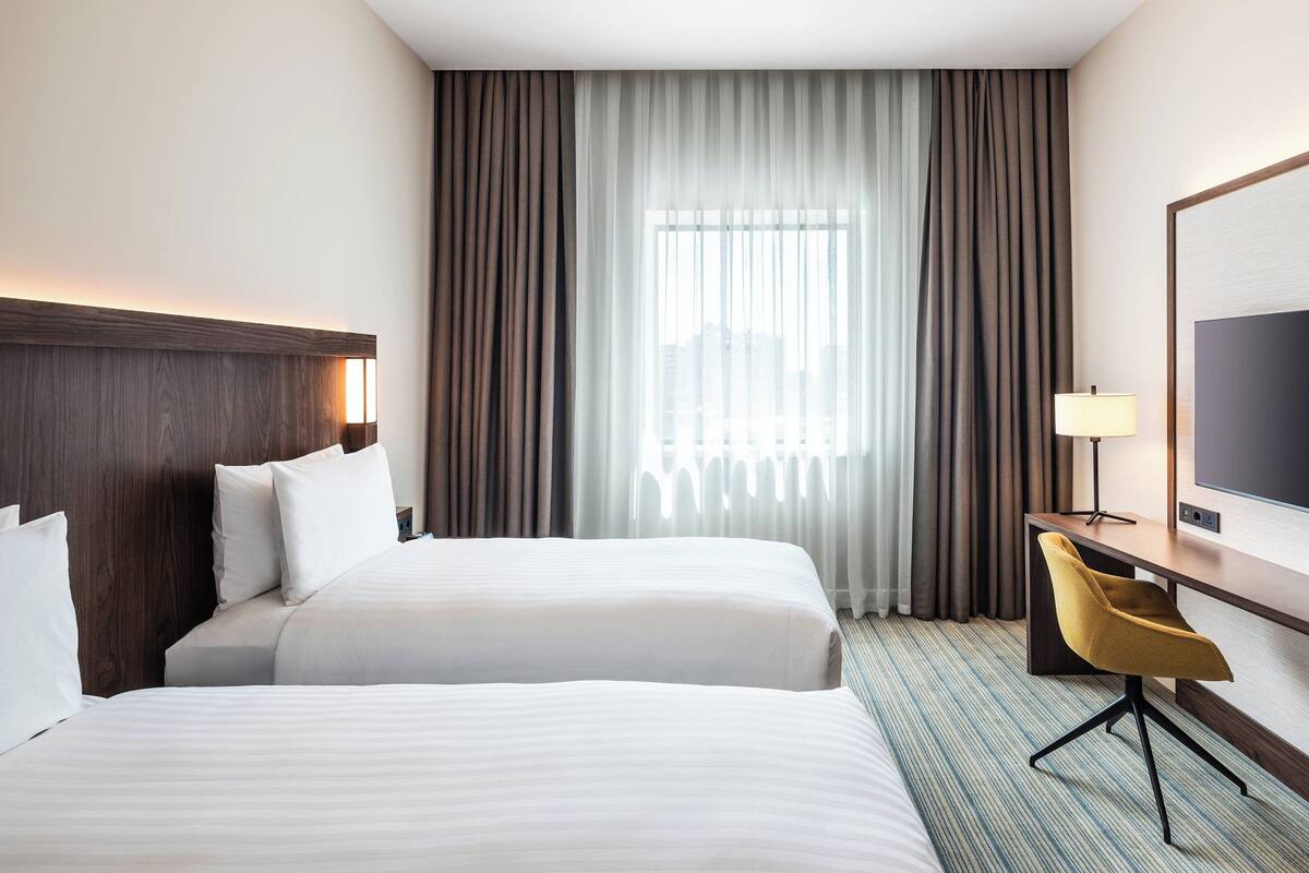 A hotel room with two beds, a desk, a chair, a TV, and a large window with curtains.