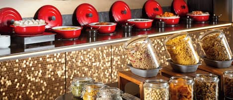 Una barra de bufet con diversos alimentos en recipientes rojos y de vidrio, dispuestos sobre una encimera con un decorativo muro de fondo.