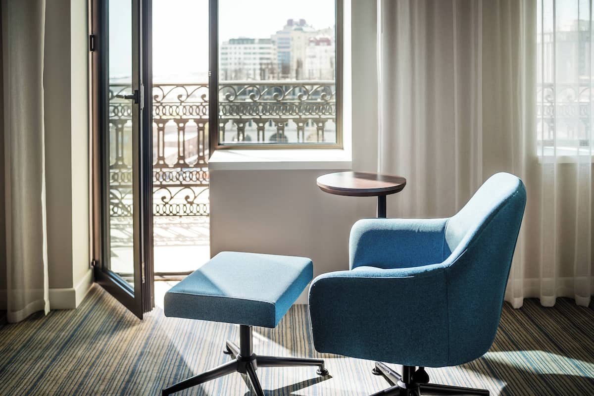 A room with a blue armchair and a small table, a balcony with a metal railing, and a view of buildings outside.