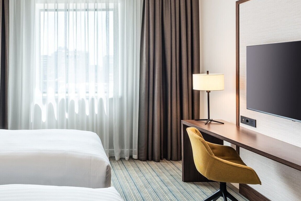 Camera d'albergo con due letti, una scrivania con una lampada, una TV e vista sulla città attraverso tende trasparenti.