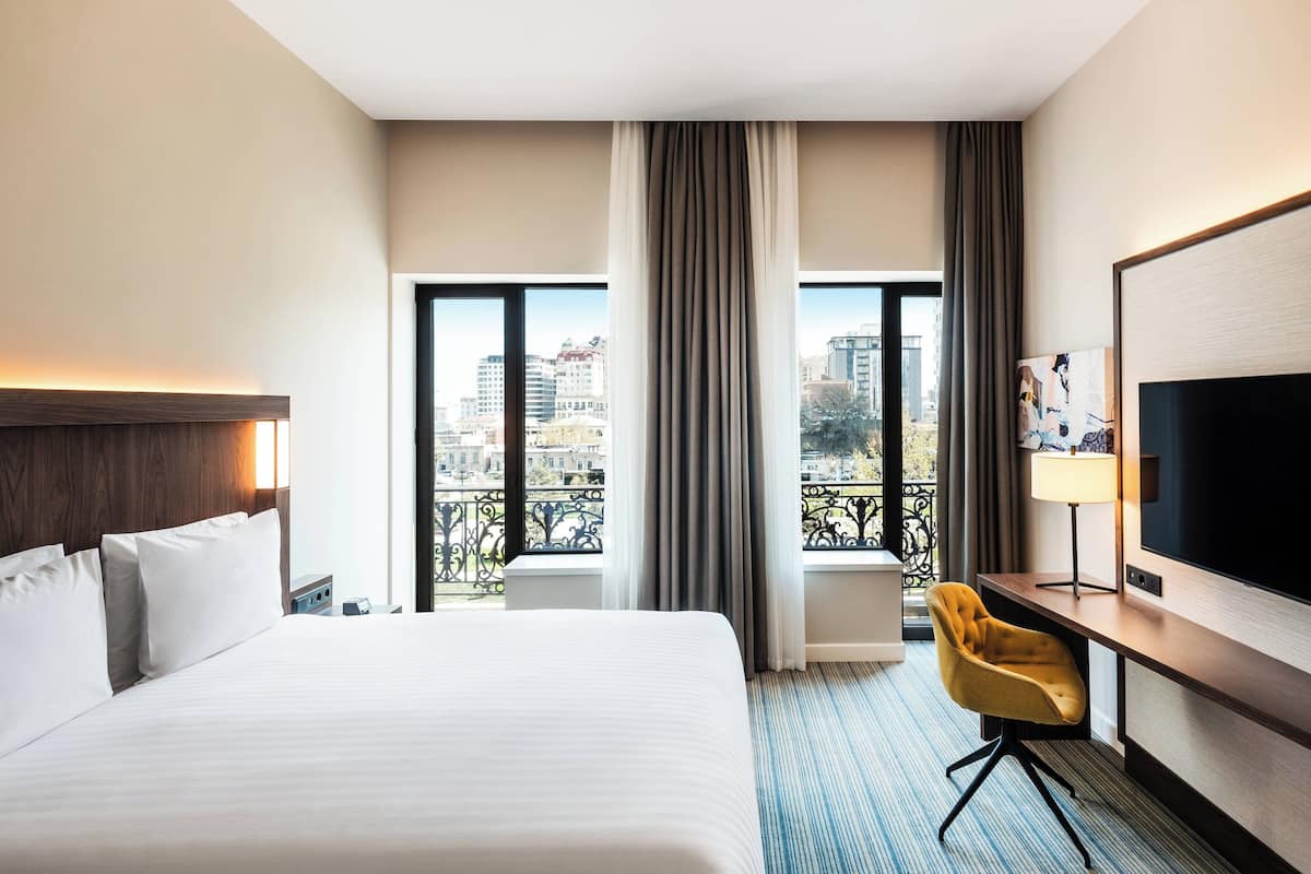 A hotel room with a large bed, a desk with a chair, a TV, and a balcony with a city view.